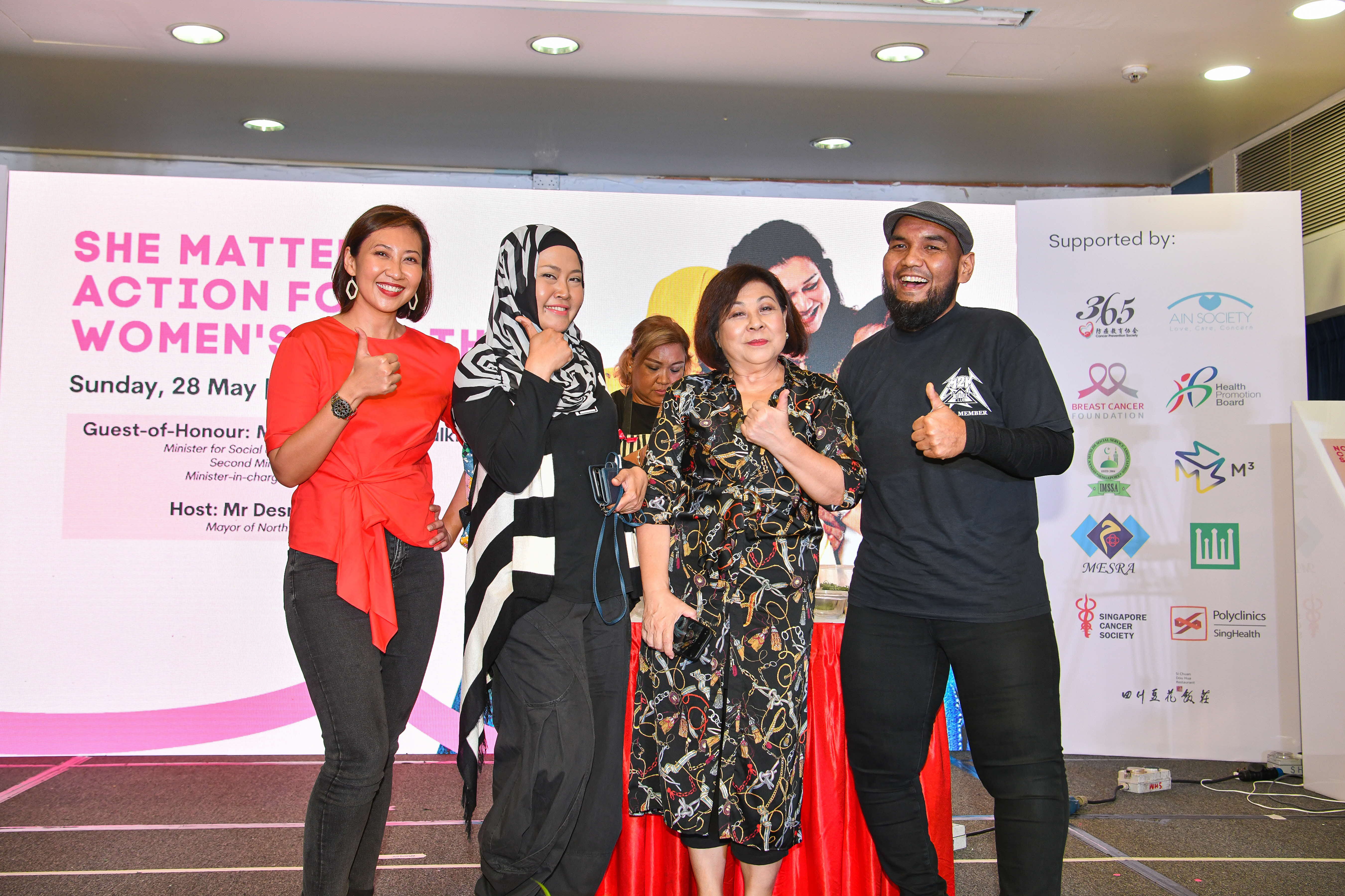 Four people pose in front of a banner and display various thumbs-up gestures; "She Matters: Action for Women's Health" event details visible.